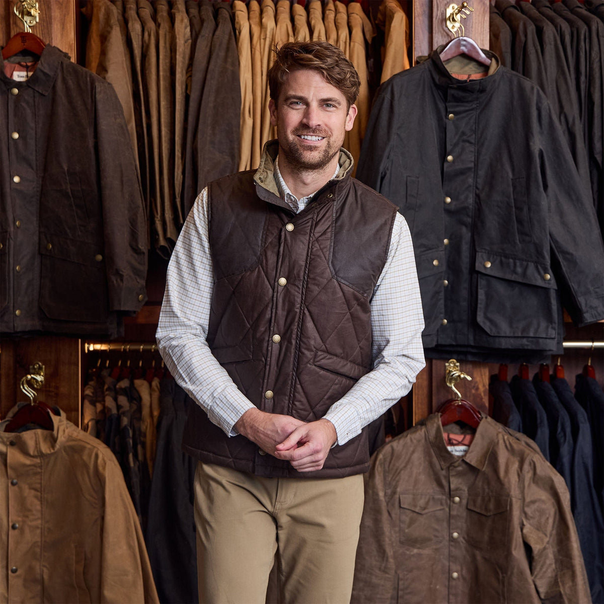 Man wears Tom Beckbe Paddock Quilted Vest over a checked shirt and khaki pants, standing by a jacket display.