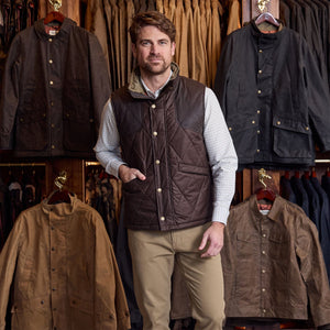 A man stands by a jacket display wearing the Tom Beckbe Paddock Quilted Vest, checked shirt, and khaki pants.