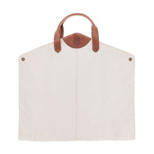 The Tom Beckbe Perdido Canvas Garment Bag features a cream rectangular body, brown leather handles, and metal snap accents.