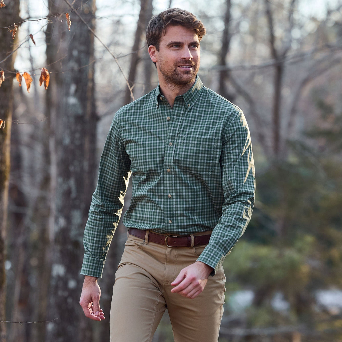 A man in a green Tom Beckbe Powell Performance Shirt and khaki pants walks outdoors in a wooded area.