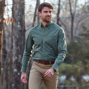 A man in a green Tom Beckbe Powell Performance Shirt and khaki pants walks outdoors in a wooded area.