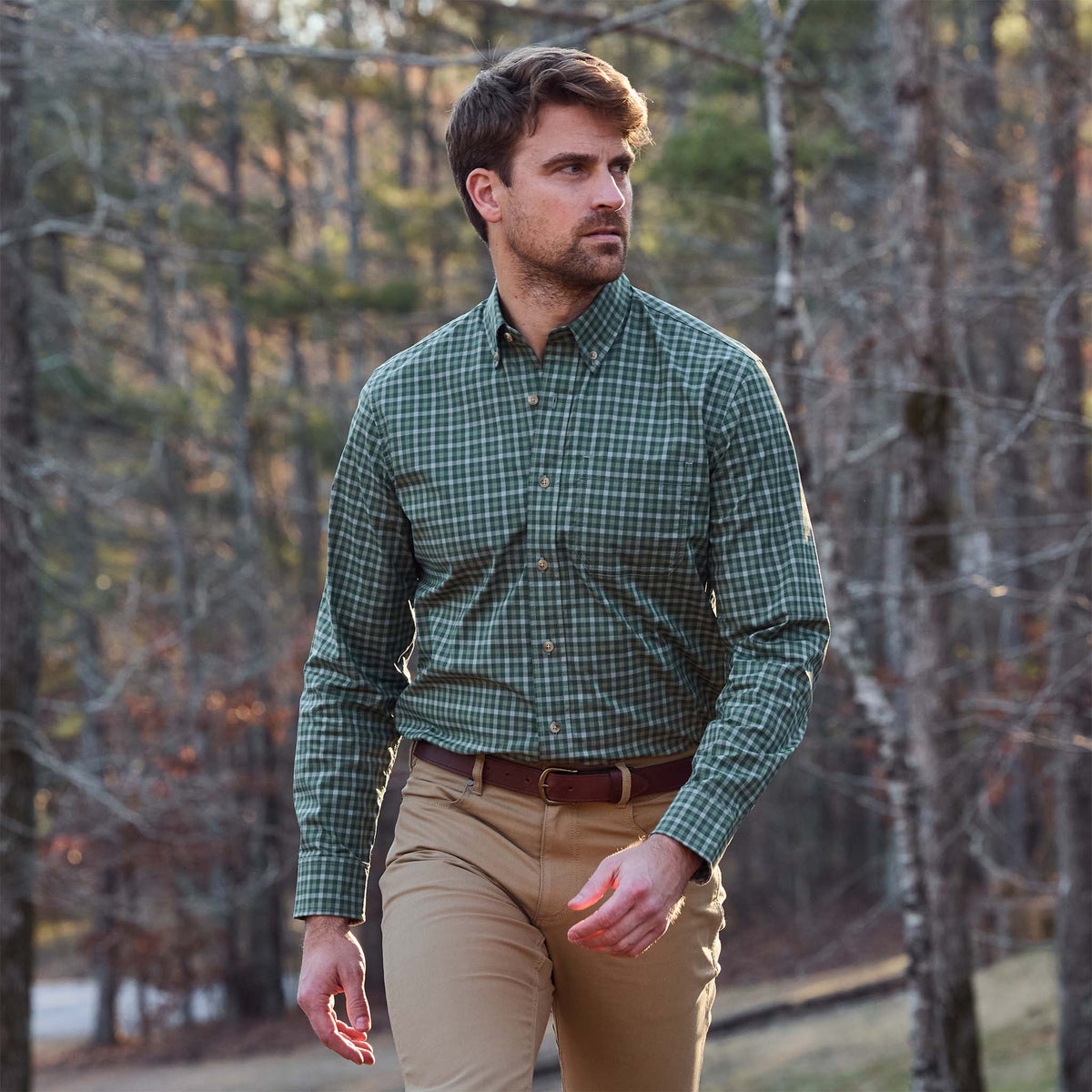 A man in a Tom Beckbe Powell Performance Shirt and khaki pants walks outdoors among bare trees.