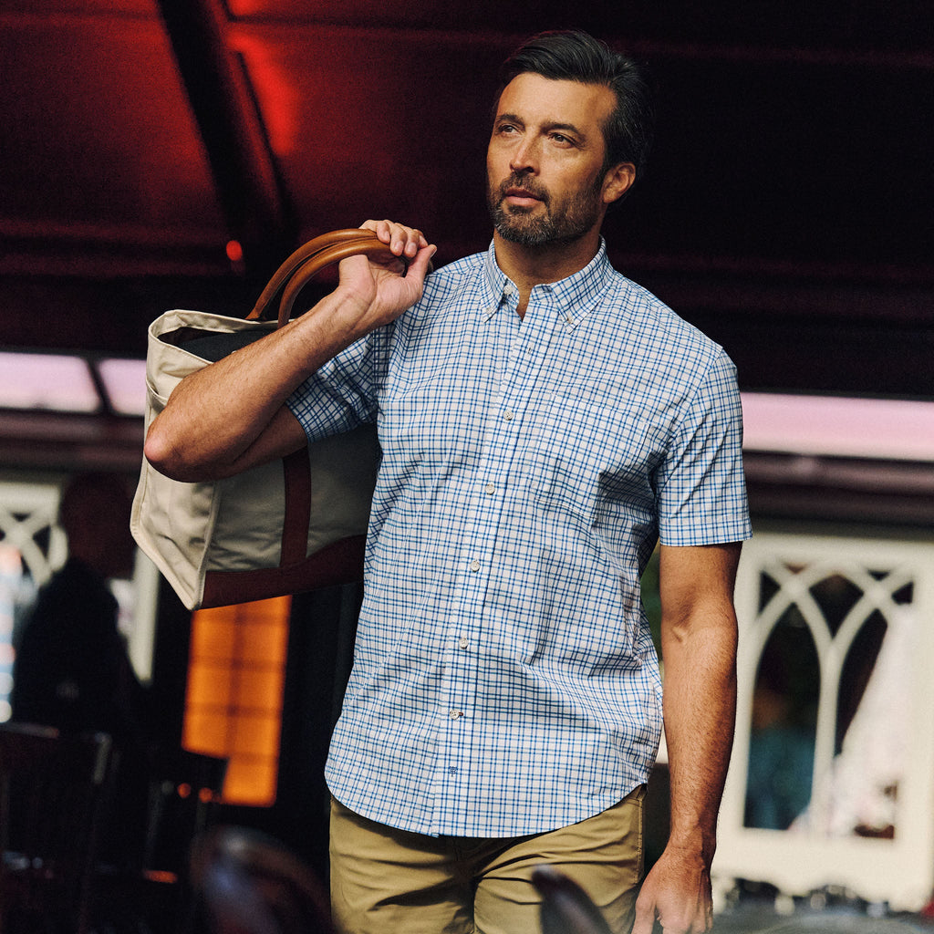 Man indoors wearing Tom Beckbe Powell Performance Short Sleeve Shirt and khaki pants, carrying a tote bag.