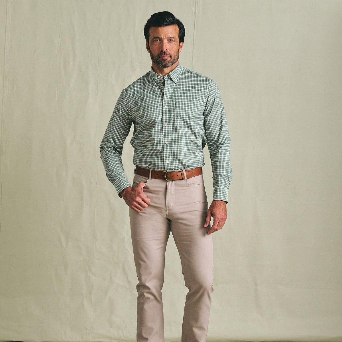 A man wears the Tom Beckbe Powell Performance Shirt, a green UPF 30+ shirt, with beige pants and a brown belt.