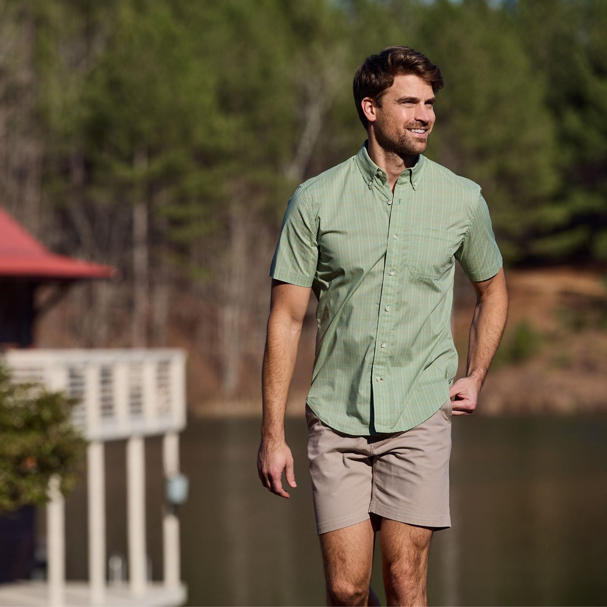 A man in a Tom Beckbe Powell Performance Shirt and khaki shorts strolls by a lake, framed by trees and a distant building.