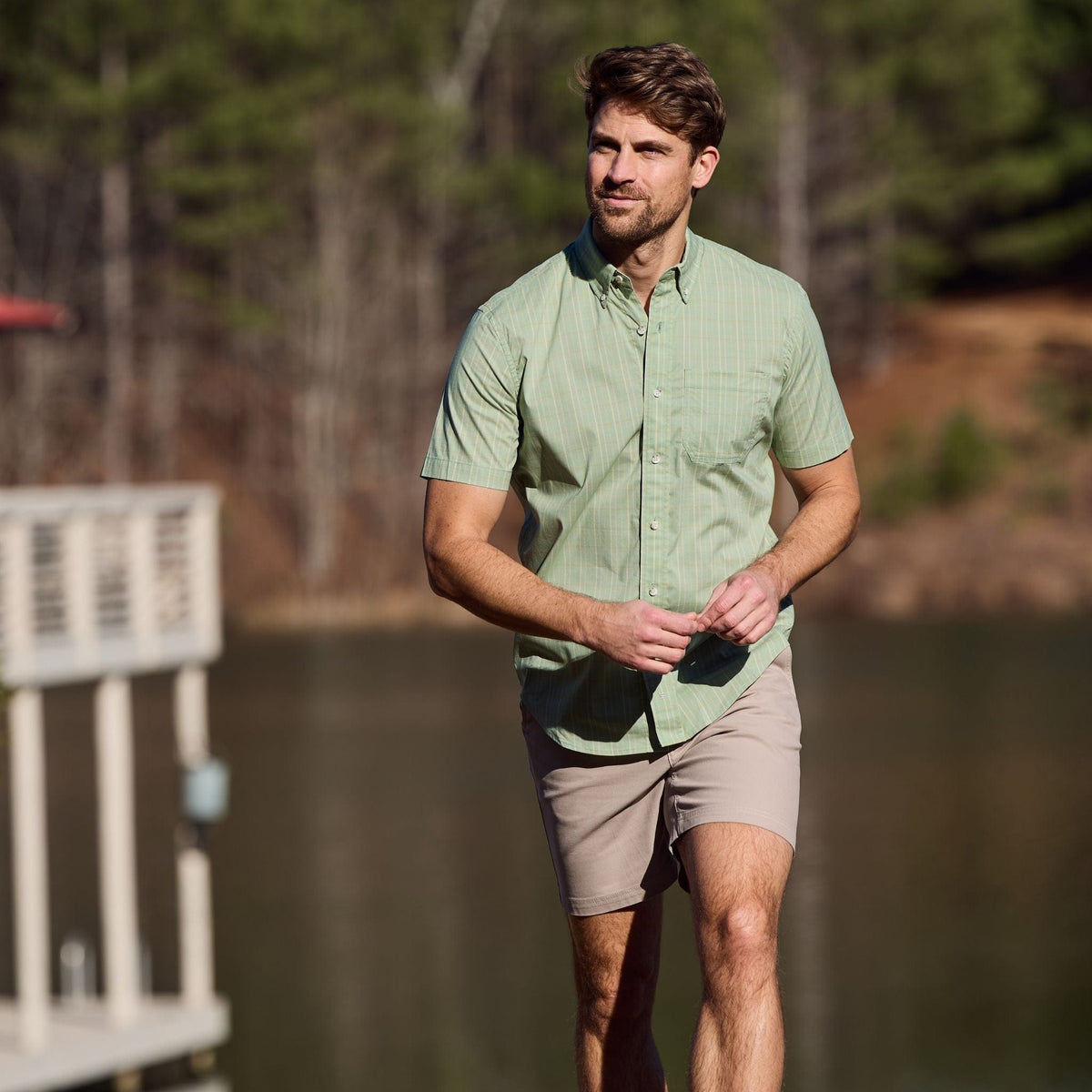 Man in a green Tom Beckbe Powell Performance Shirt and beige shorts walks near a lake and trees, enjoying the outdoors.