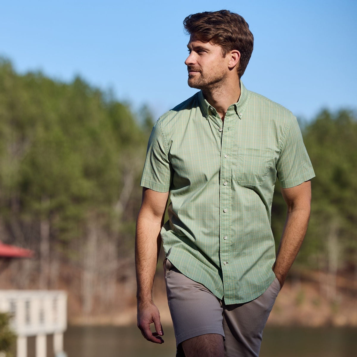 A man wears a Tom Beckbe Powell Performance Shirt and khaki shorts, walking by a lake on a sunny day.