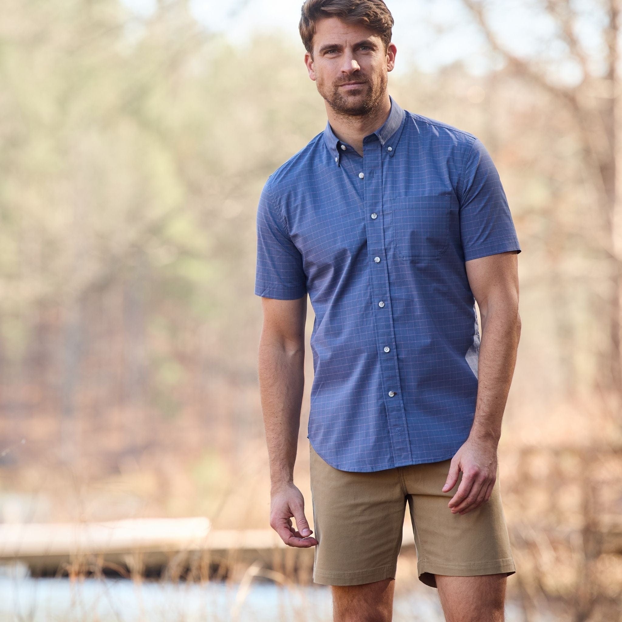 A man in a blue Tom Beckbe Powell Performance Shirt and khaki shorts stands outside with trees and water behind him.