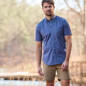A man in a blue Tom Beckbe Powell Performance Shirt and khaki shorts stands outside with trees and water behind him.