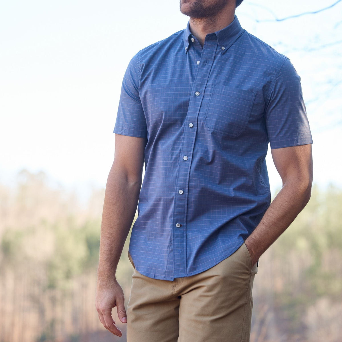 Man in a Tom Beckbe Powell Performance Shirt stands outdoors with khaki pants against a blurred forest backdrop.