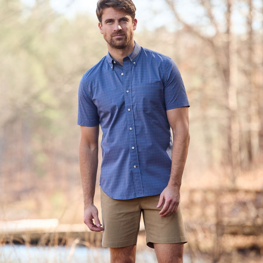Man outdoors in a blue shirt and Tom Beckbe Reid short, with trees and a wooden bridge in the background.