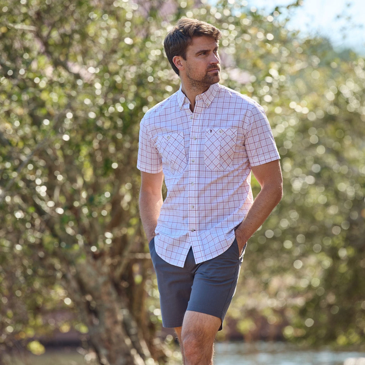 A man outdoors in Tom Beckbes Reid Short and a plaid shirt, gazes aside with trees behind.