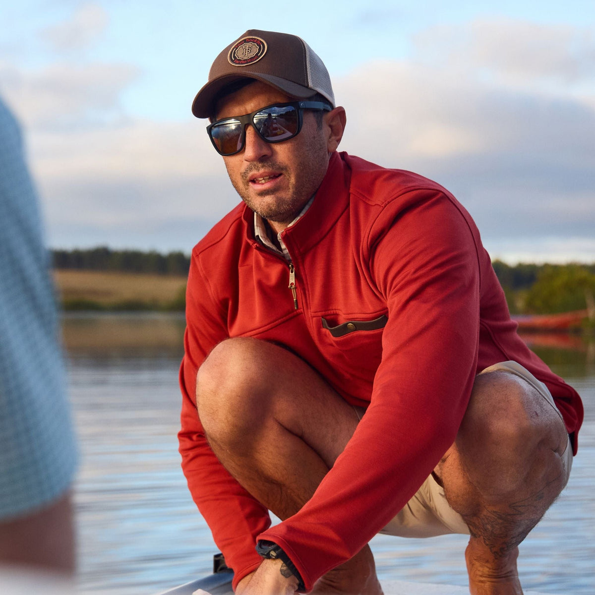 A man squats near water in a cap and shorts, with a Tom Beckbe Tucker Fleece Quarter Zip in the background.