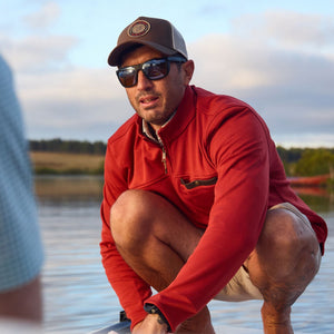 A man squats near water in a cap and shorts, with a Tom Beckbe Tucker Fleece Quarter Zip in the background.