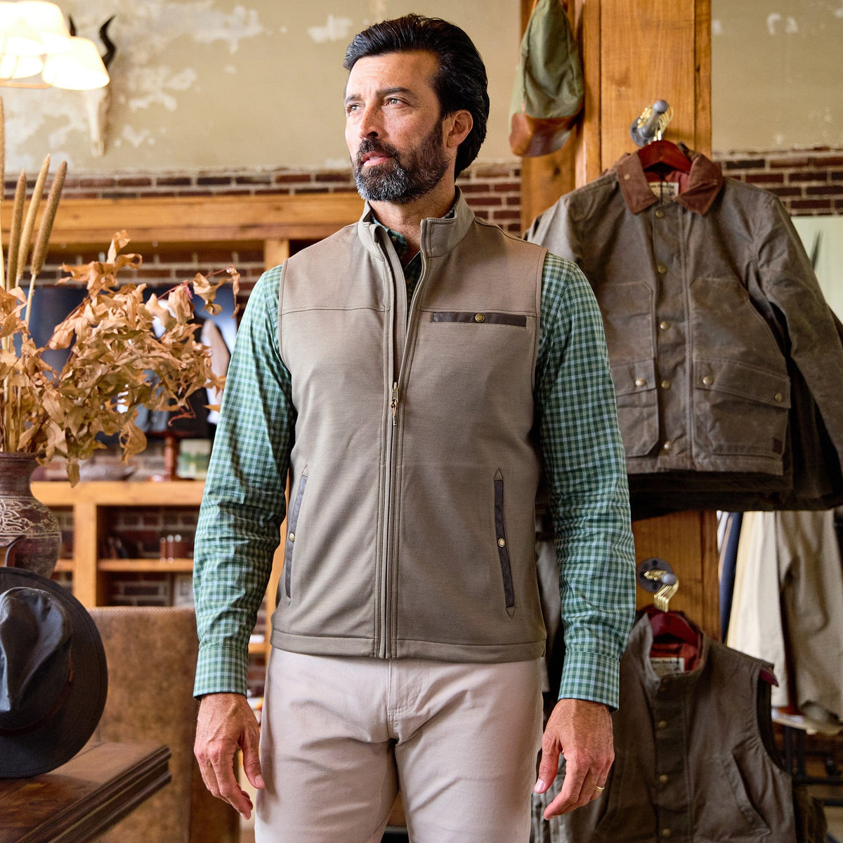 A man in a green checkered shirt and Tom Beckbe Tucker Fleece Vest stands indoors near jackets and rustic decor.