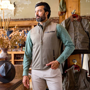 A man in a green plaid shirt and Tom Beckbe Tucker Fleece Vest stands in a rustic store with jackets and hats.