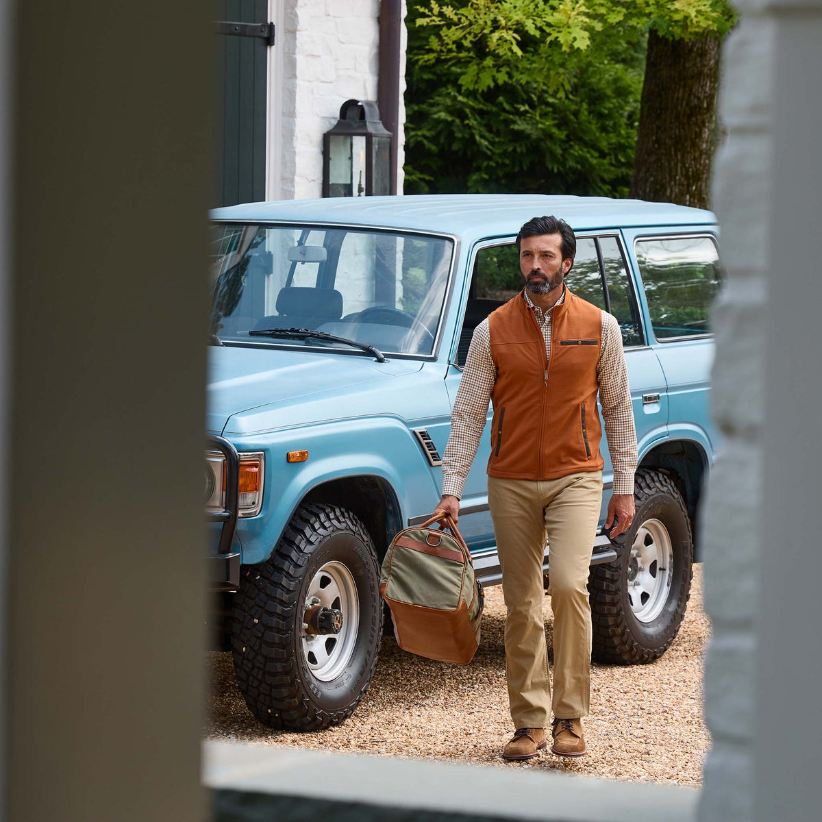 A man in a Tom Beckbe Tucker Fleece Vest carries a duffel bag by a blue SUV on a gravel drive near a white building.
