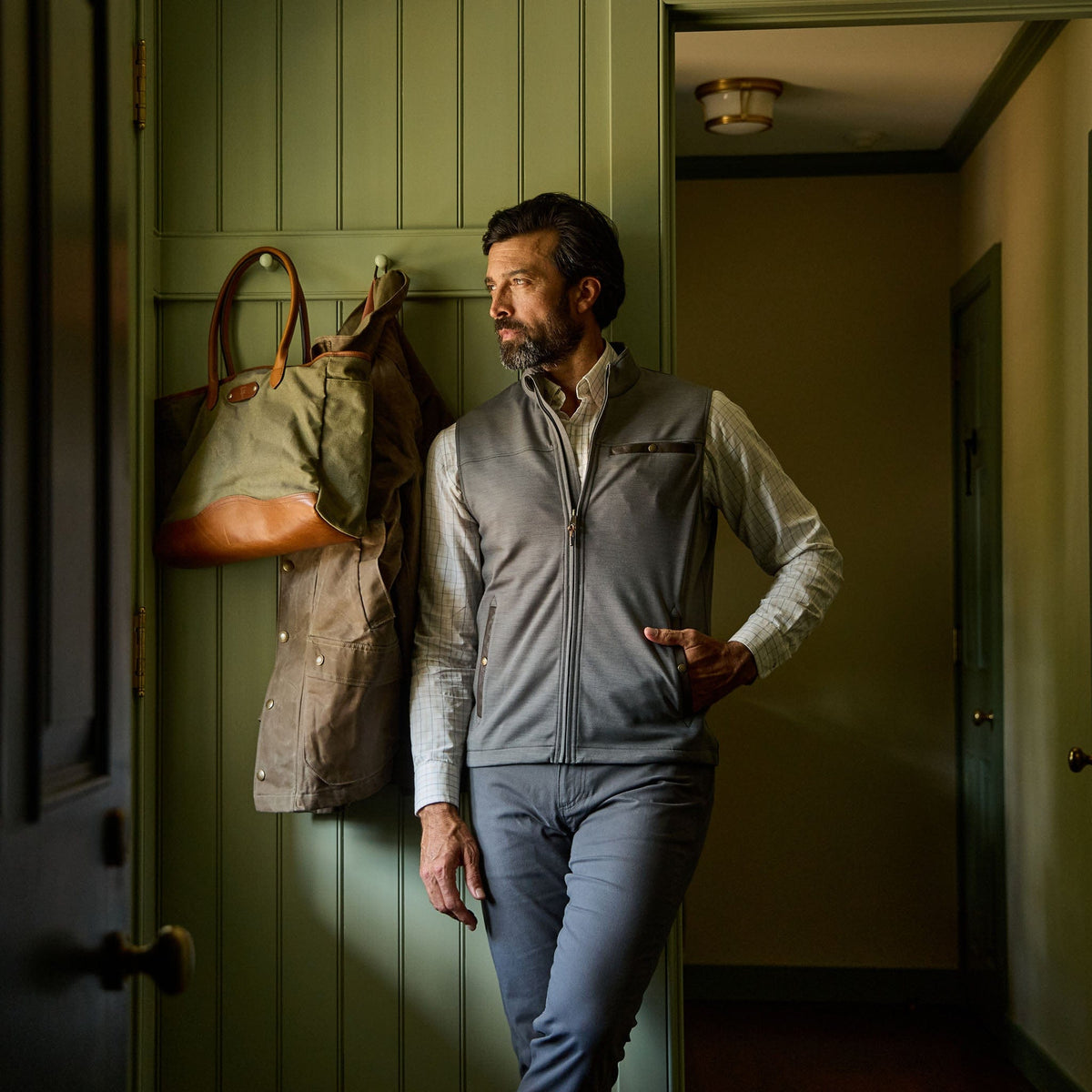 A man in a Tom Beckbe Tucker Fleece Vest stands indoors by a green wall with coats and a bag hanging behind him.