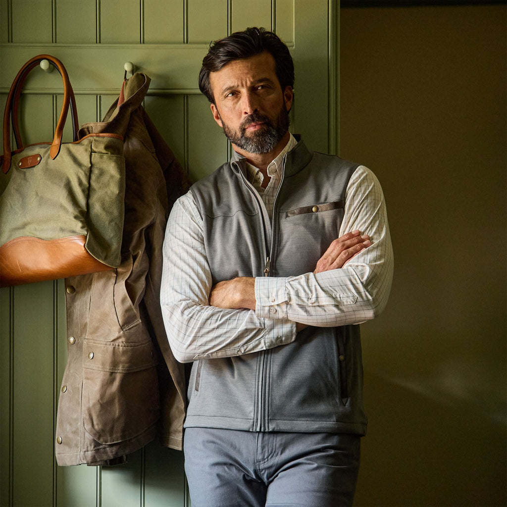 A man wears the Tom Beckbe Tucker Fleece Vest indoors by a green wall with a coat and bag hanging on hooks.