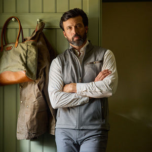 A man wears the Tom Beckbe Tucker Fleece Vest indoors by a green wall with a coat and bag hanging on hooks.