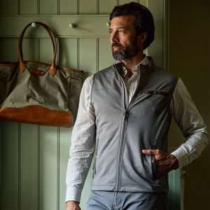 Man wears Tom Beckbe Tucker Fleece Vest over plaid shirt, standing indoors beside a green wall and leather bag.