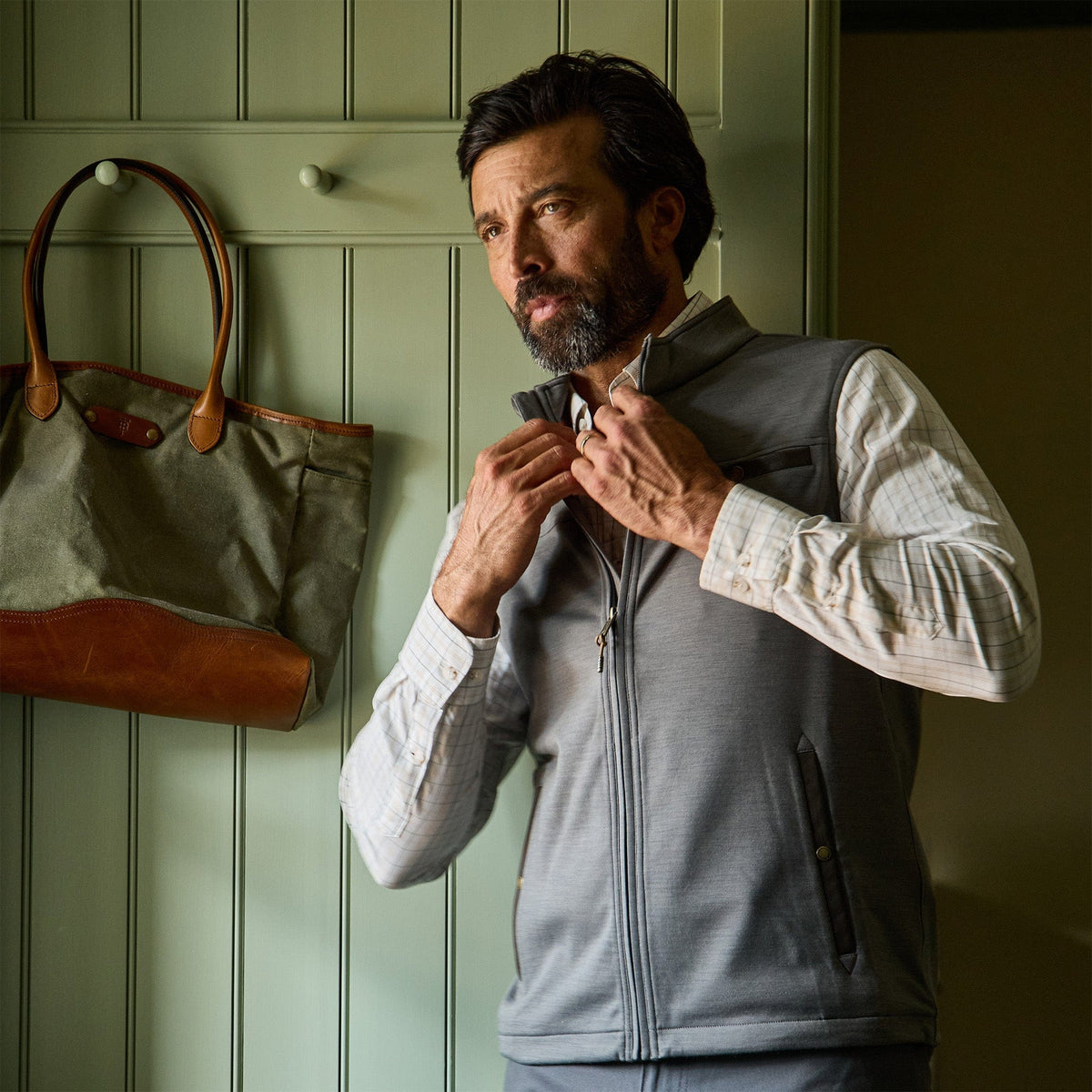 Man wearing a Tom Beckbe Tucker Fleece Vest over a plaid shirt zips up indoors beside a tote bag on a hook.