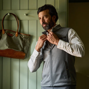 Man wearing a Tom Beckbe Tucker Fleece Vest over a plaid shirt zips up indoors beside a tote bag on a hook.