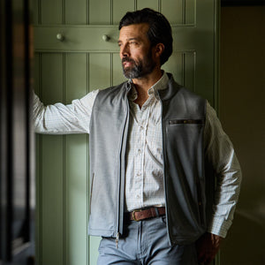 Man with dark hair and beard in a Tom Beckbe Tucker Fleece Vest stands by a green paneled wall, gazing outside.