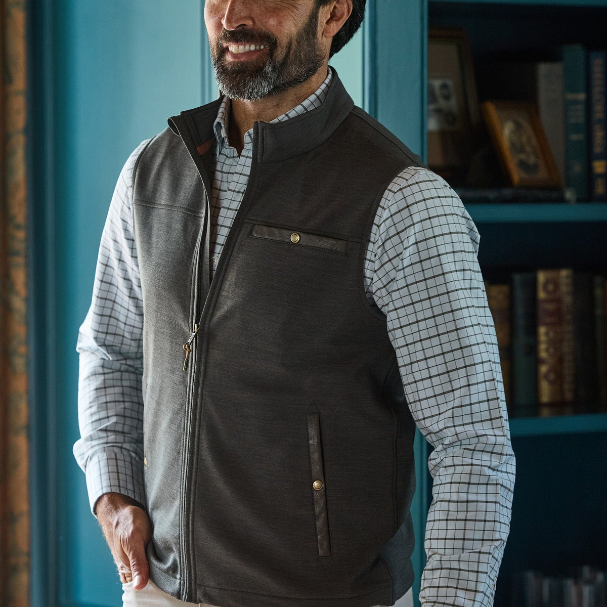 Man wears Tom Beckbe Tucker Fleece Vest over a checkered shirt, standing indoors by a blue bookshelf, smiling hands in pockets.