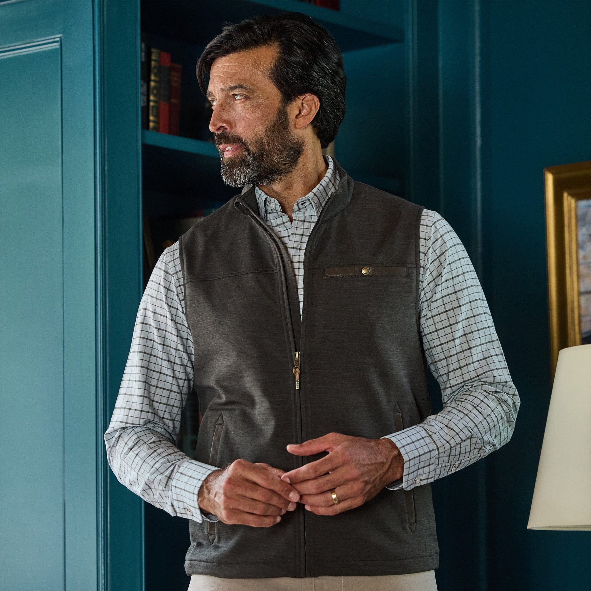 A man in a checked shirt and Tom Beckbe Tucker Fleece Vest stands indoors by a blue wall and bookshelf, looking aside.