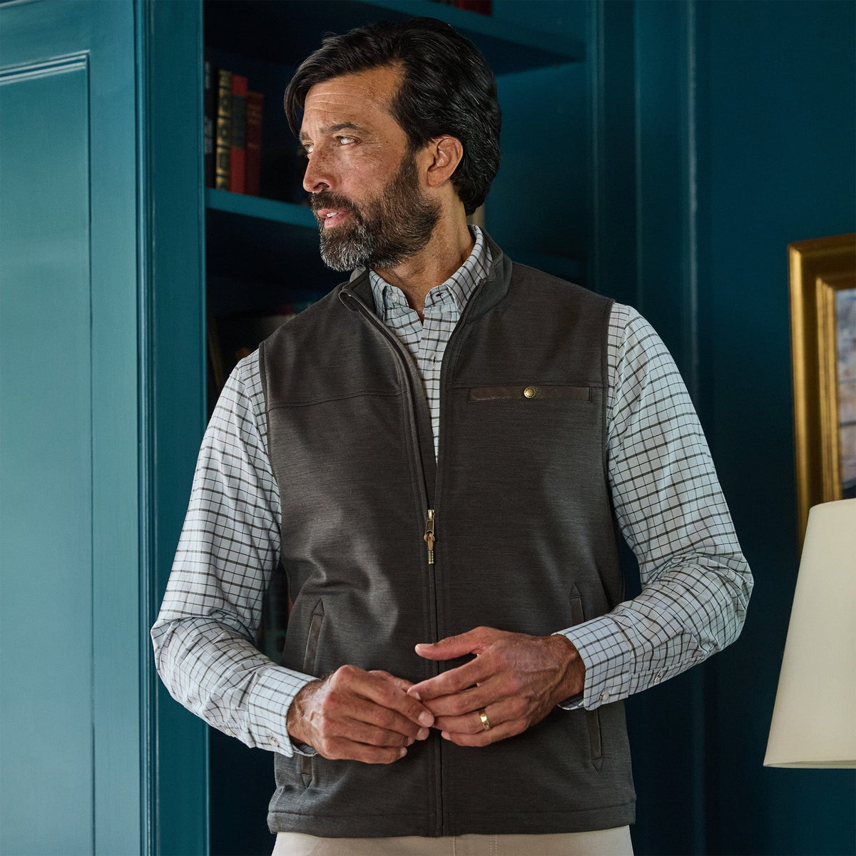 A man in a checked shirt and Tom Beckbe Tucker Fleece Vest stands indoors by a blue wall and bookshelf, looking aside.