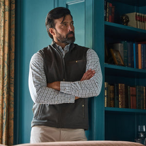 Bearded man in a Tom Beckbe Tucker Fleece Vest stands with arms crossed before a blue bookshelf.