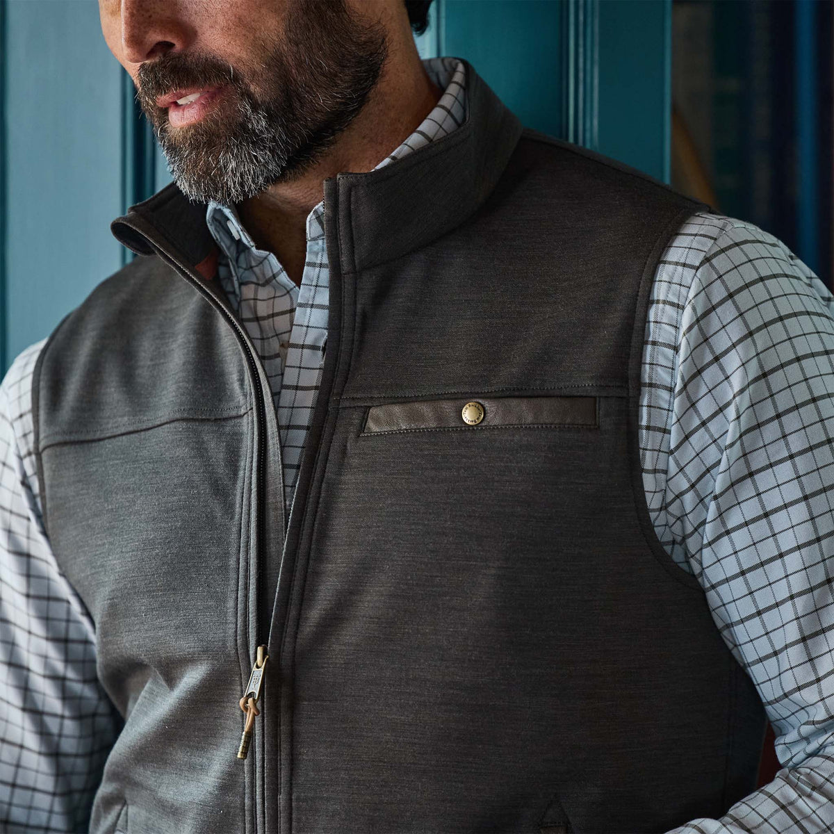 Man in Tom Beckbe Tucker Fleece Vest and checkered shirt stands indoors by a blue wall.