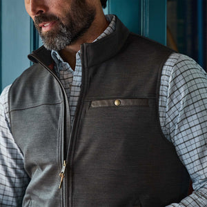 Man in Tom Beckbe Tucker Fleece Vest and checkered shirt stands indoors by a blue wall.