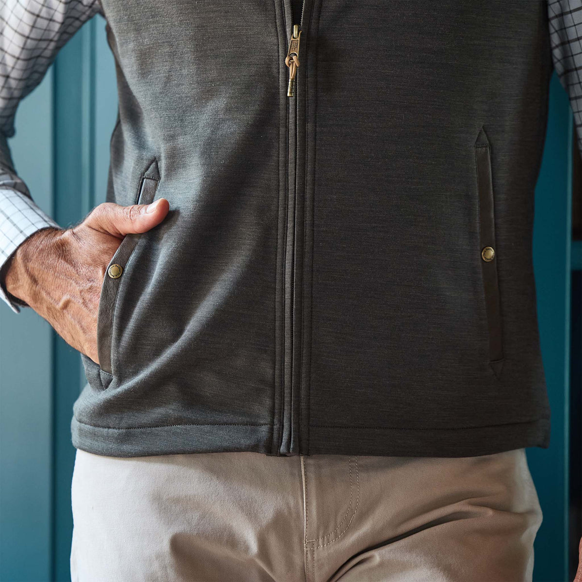 A person wearing the Tom Beckbe Tucker Fleece Vest, a checked shirt, and beige pants with one hand in the vest pocket.