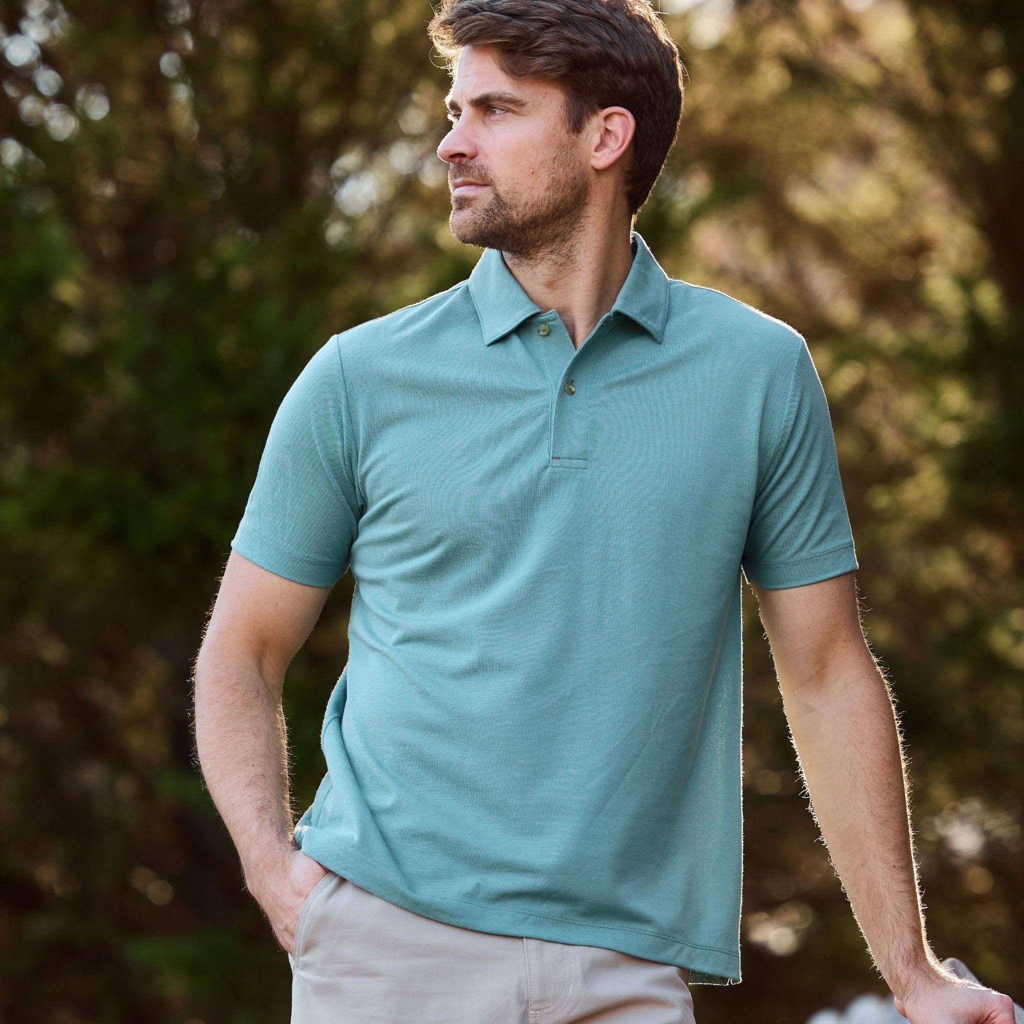 A man outdoors looks left, wearing a Tom Beckbe Coastal Polo Shirt against a backdrop of trees.