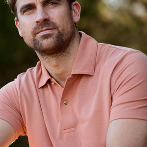 A bearded man in a salmon-colored Tom Beckbe Coastal Polo Shirt relaxes outdoors under the sunny sky.