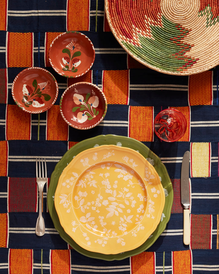 Yellow floral dessert plate on a green charger on a colorful woven tablecloth with fork, knife, red glass, decorative bowls and woven basket