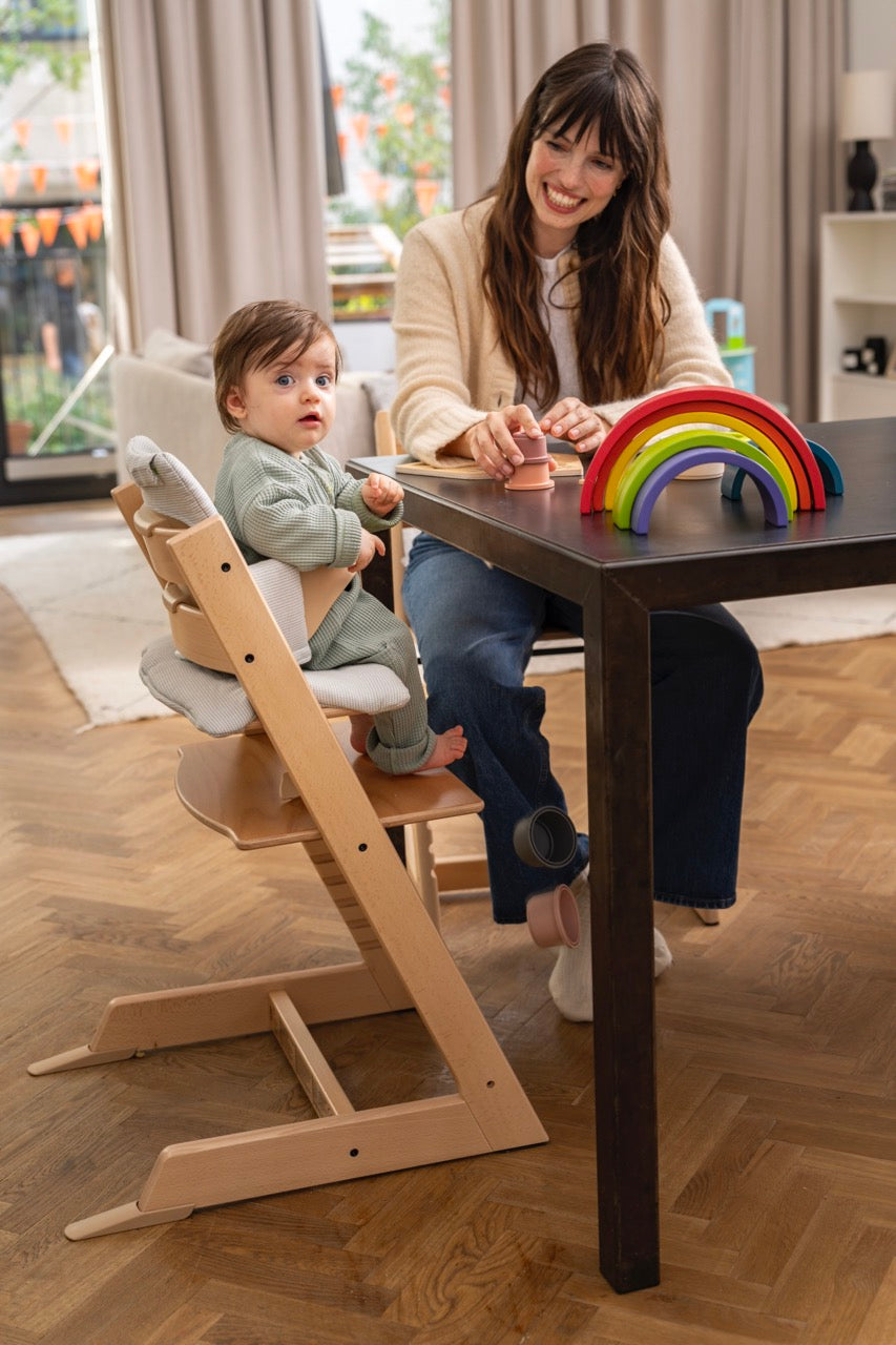 Baby sitting in Tripp Trapp Baby Set in Natural at a dining table while a smiling woman plays with wooden rainbow toys