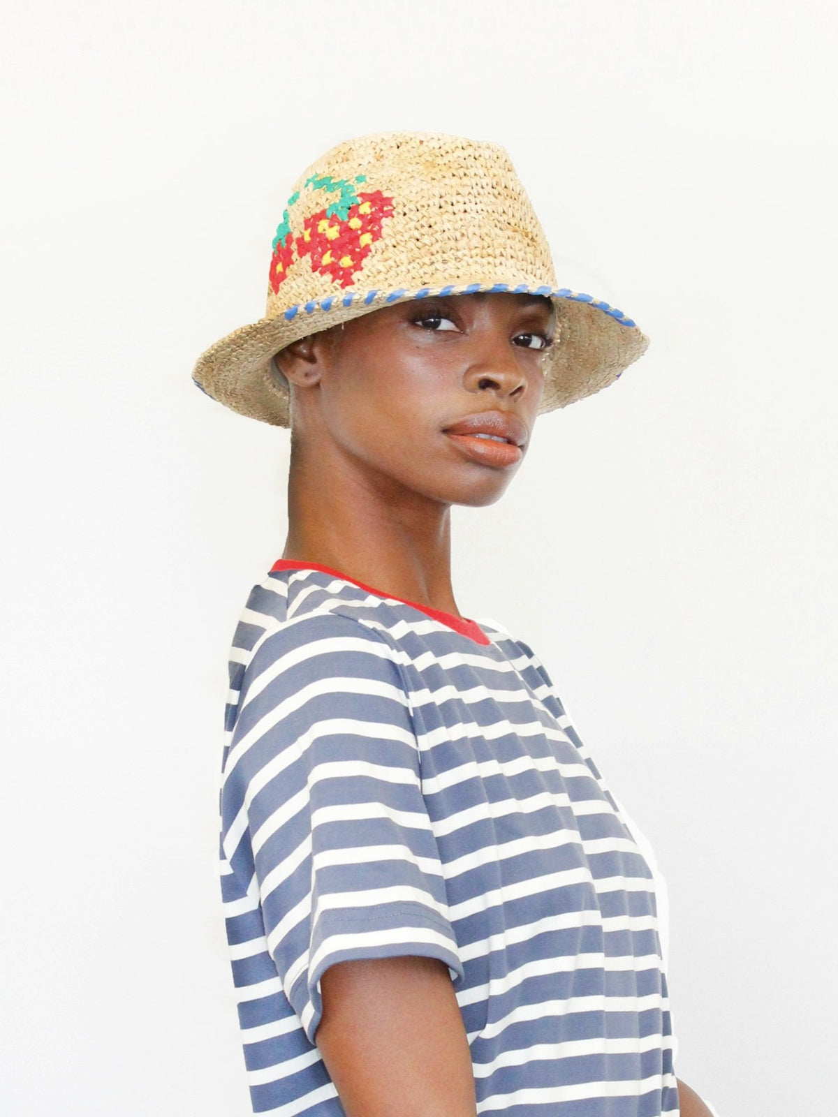 Side view of Tutti Frutti Hat on model showcasing straw crochet with embroidered strawberry motif and blue braided brim against white background