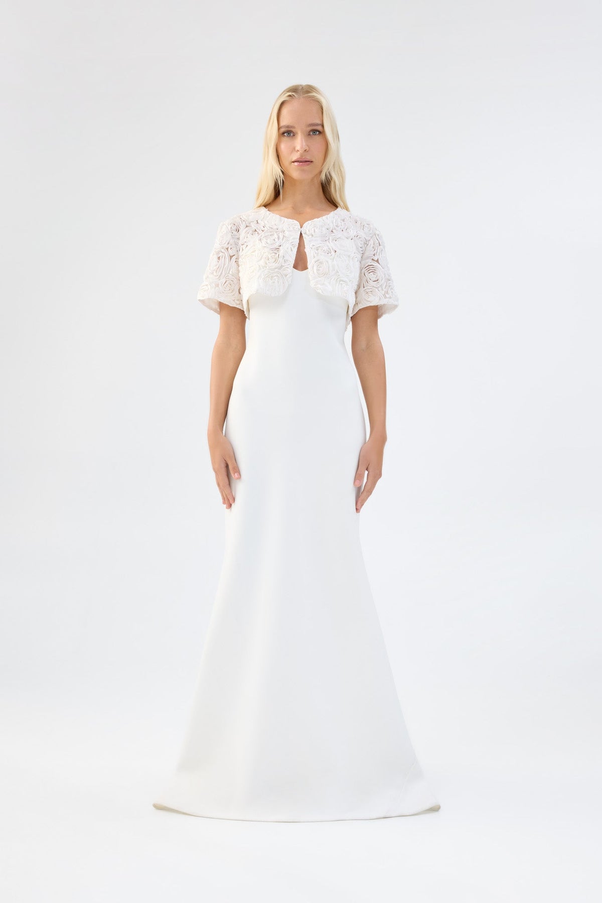 A model viewed from the front wearing a white floral textured bridal bolero over a white gown.
