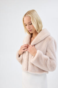 Close-up of a model wearing a blush pink faux fur jacket with notch lapel and cropped fit, hands adjusting the collar