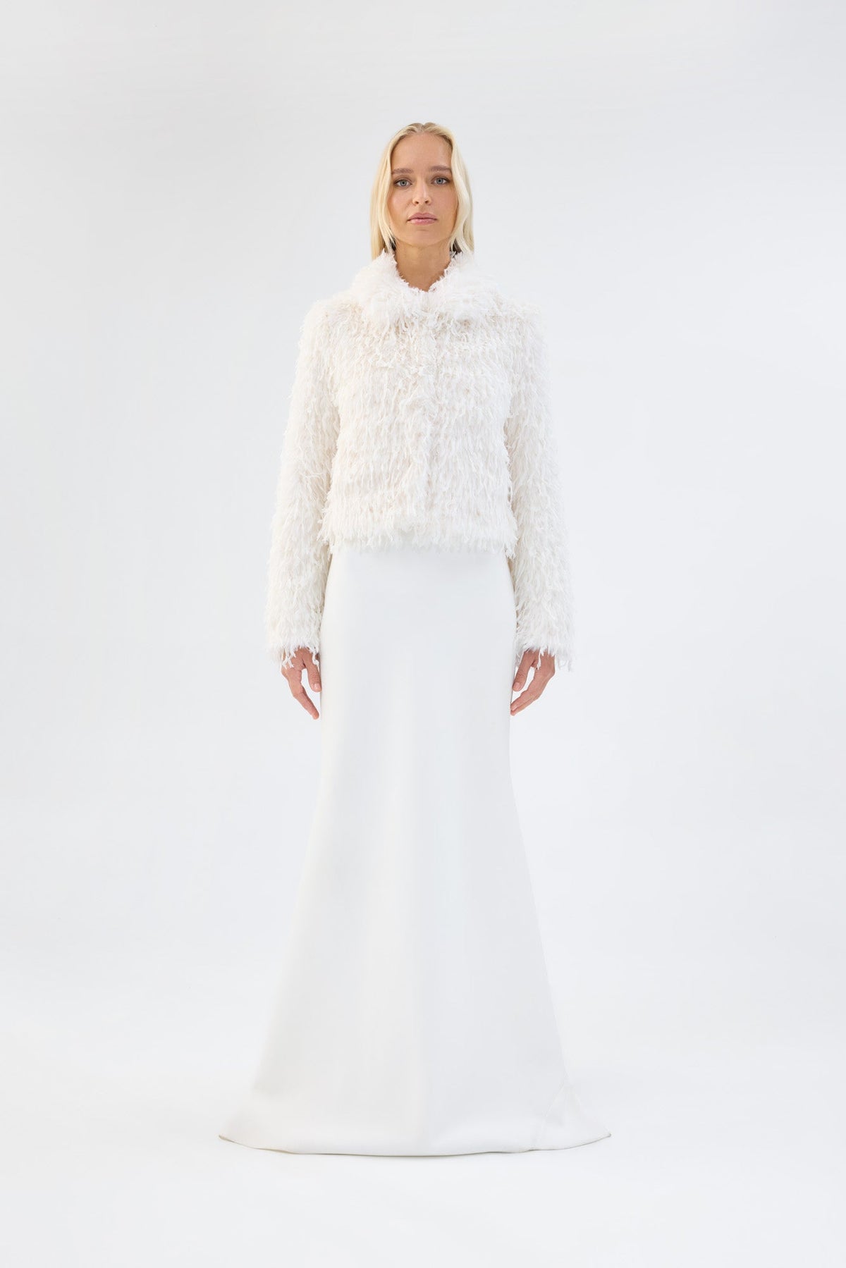 A model viewed from the front wearing a white textured faux fur jacket over a white gown.
