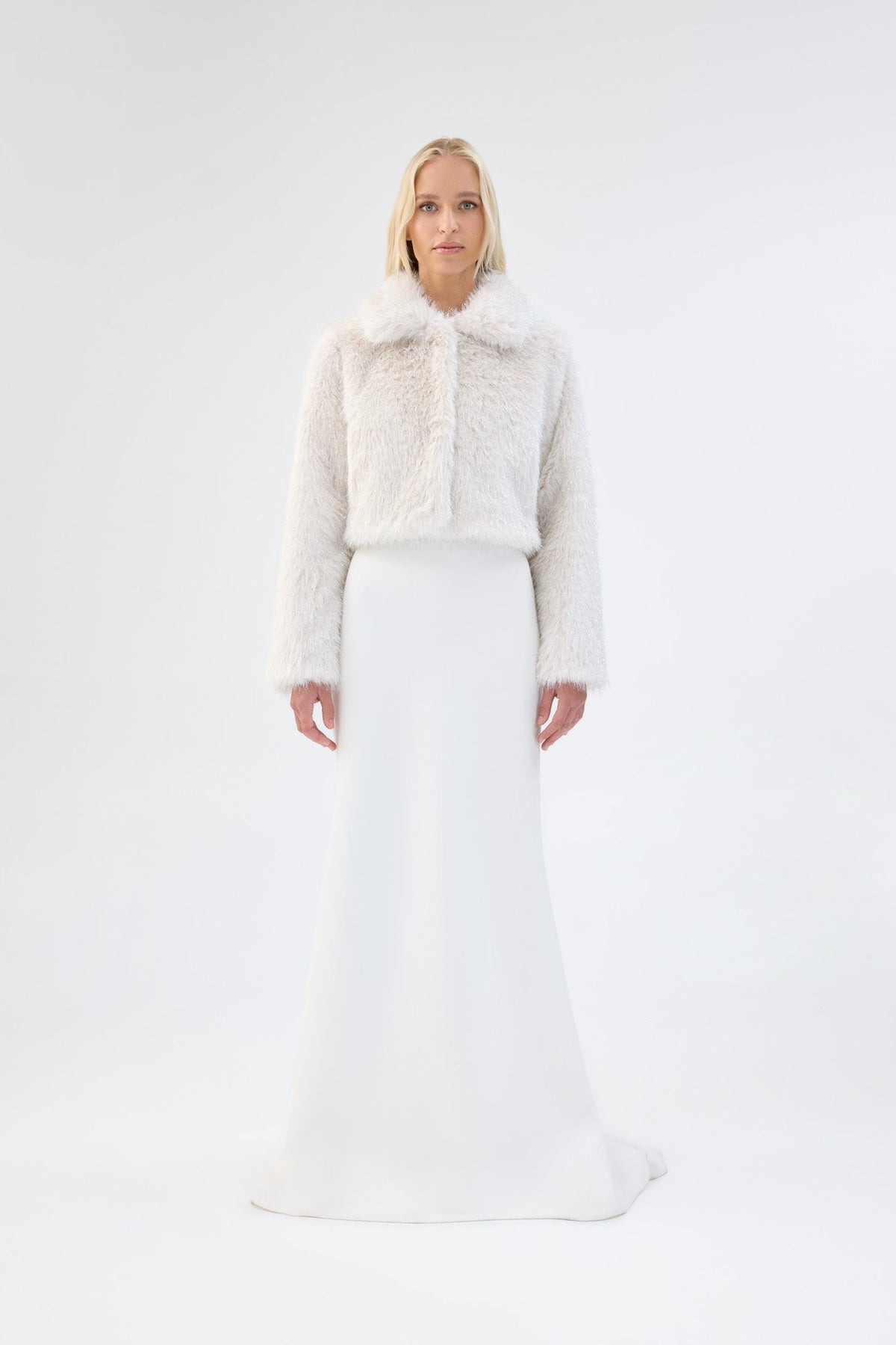 A model viewed from the front wearing a white cropped faux fur jacket over a white gown.