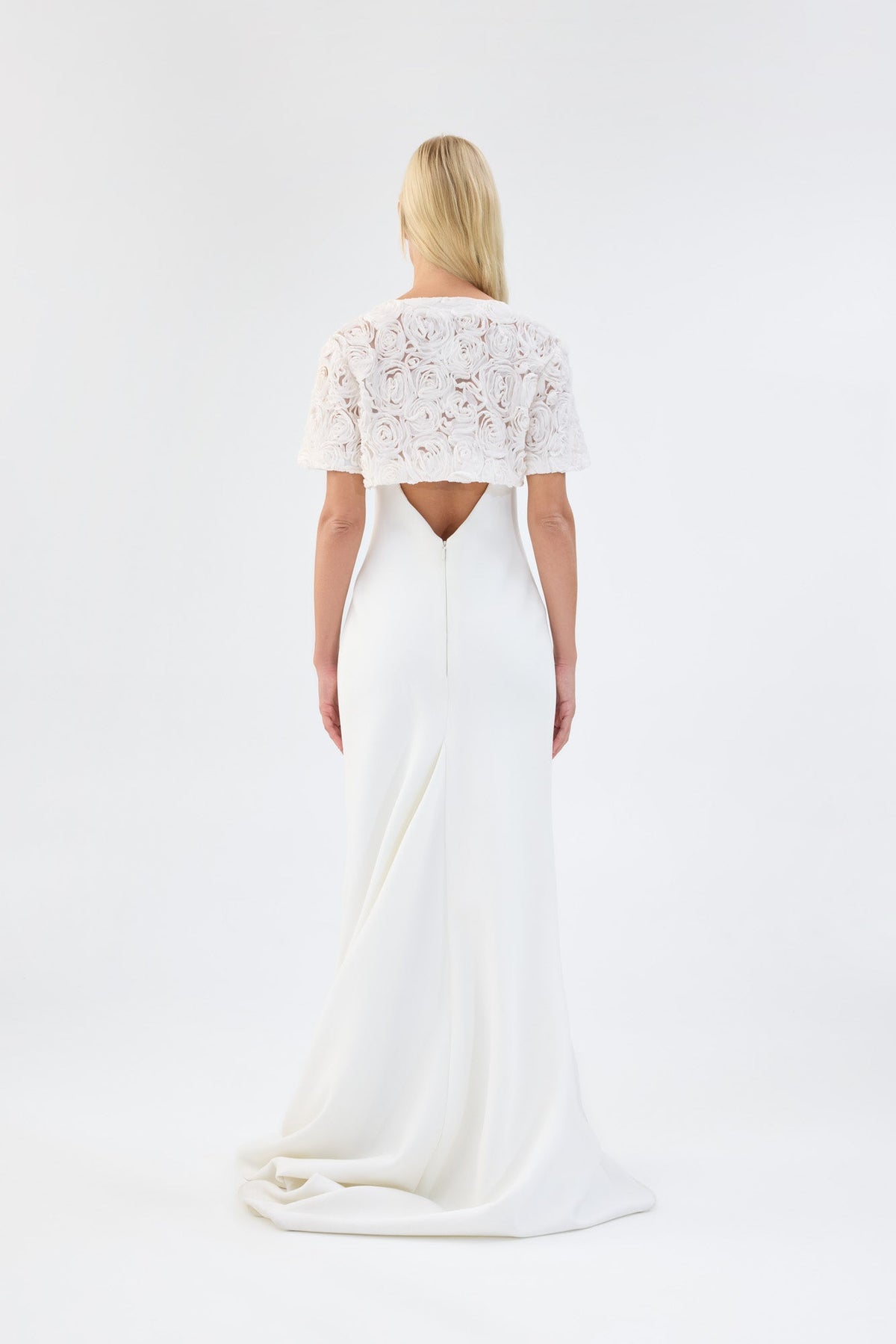 A model viewed from the back wearing a white floral textured bridal bolero over a white gown.