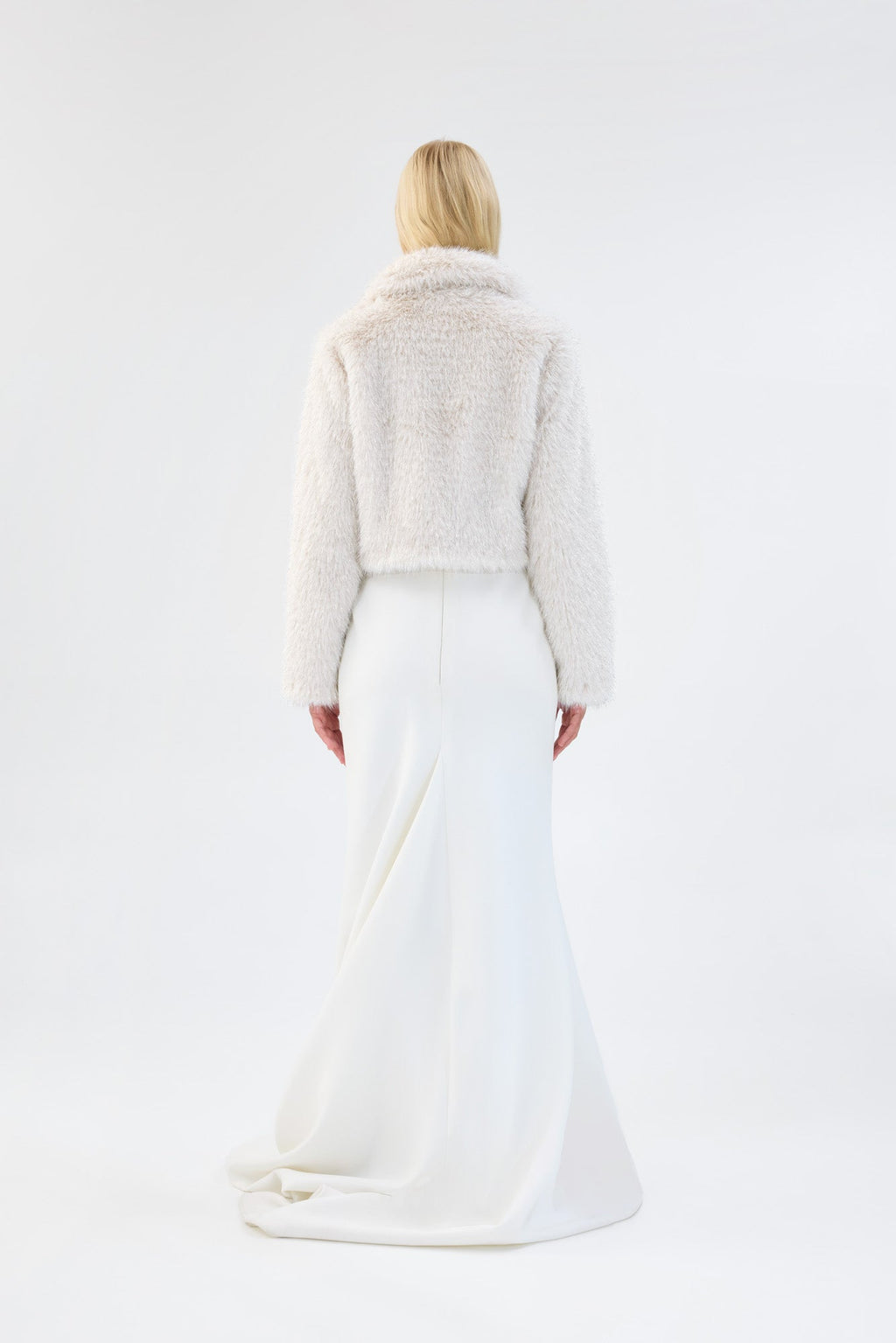 A model viewed from the back wearing a white cropped faux fur jacket over a white gown.