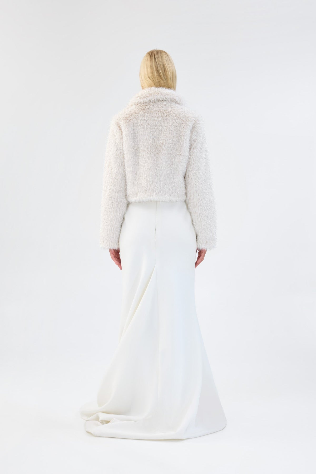 A model viewed from the back wearing a white cropped faux fur jacket over a white gown.