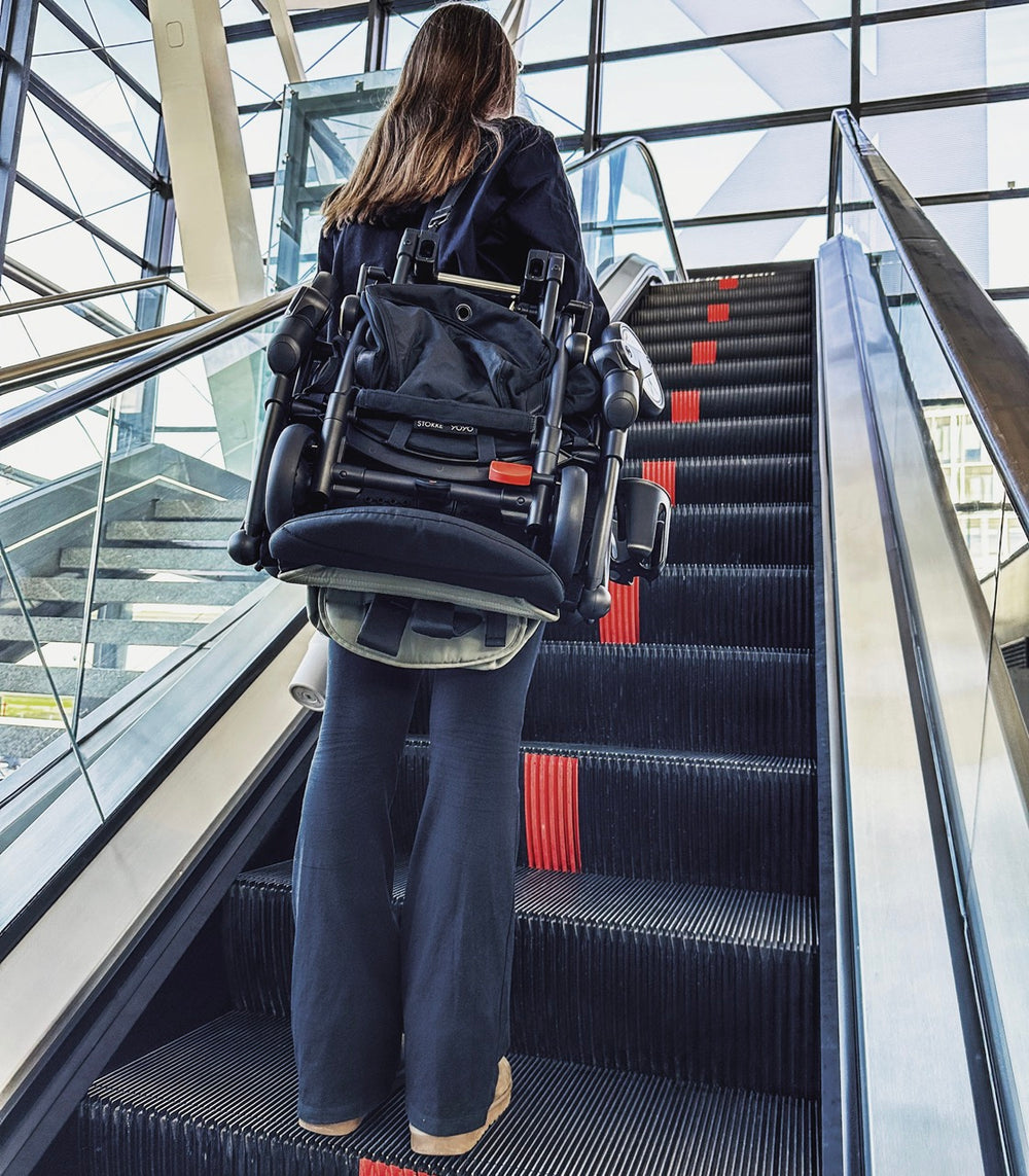 Woman carrying folded olive YOYO3 stroller up an escalator in a glass-walled terminal