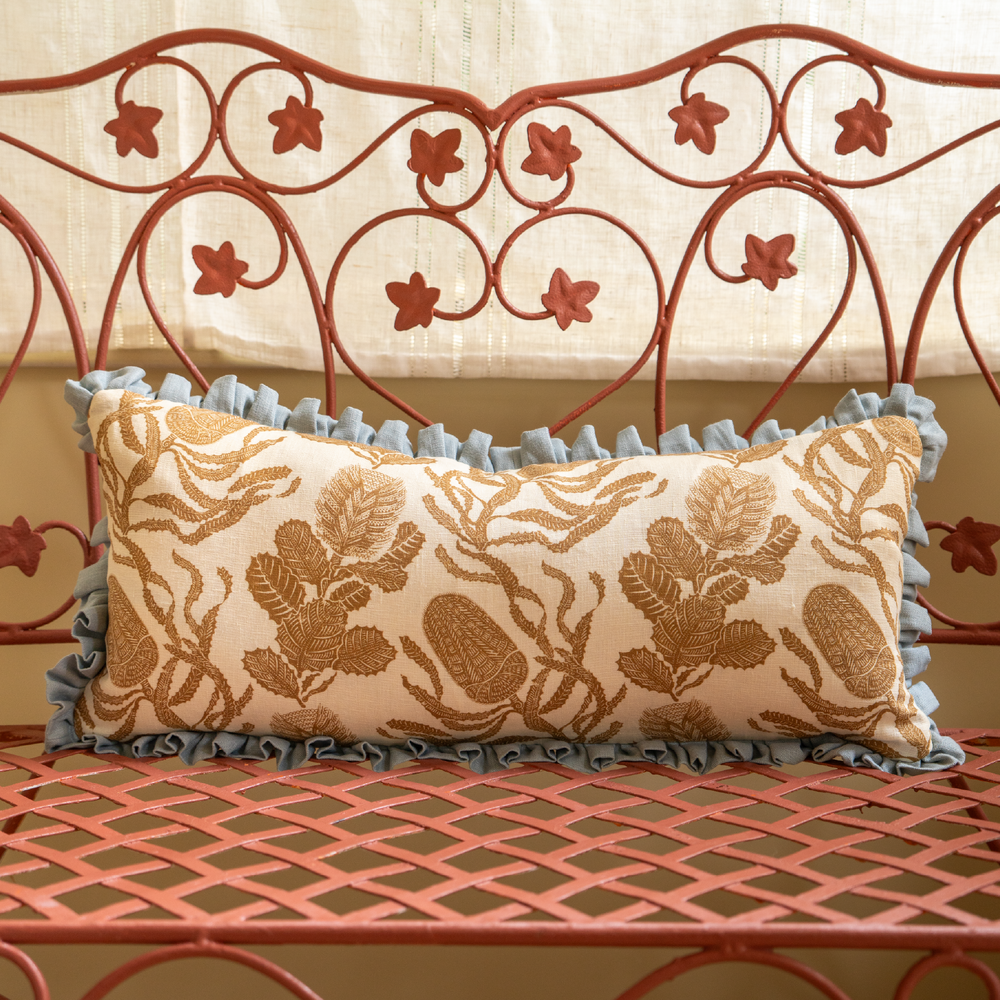 Front view of Cape Botanical Print Cushion in Old Gold showcasing botanical print and Porcelain Blue frill on an iron bench