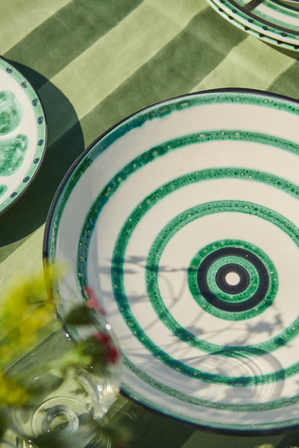 Top-down view of Bertie serving bowl showcasing hand-painted green concentric rings on white ceramic with green striped tablecloth background